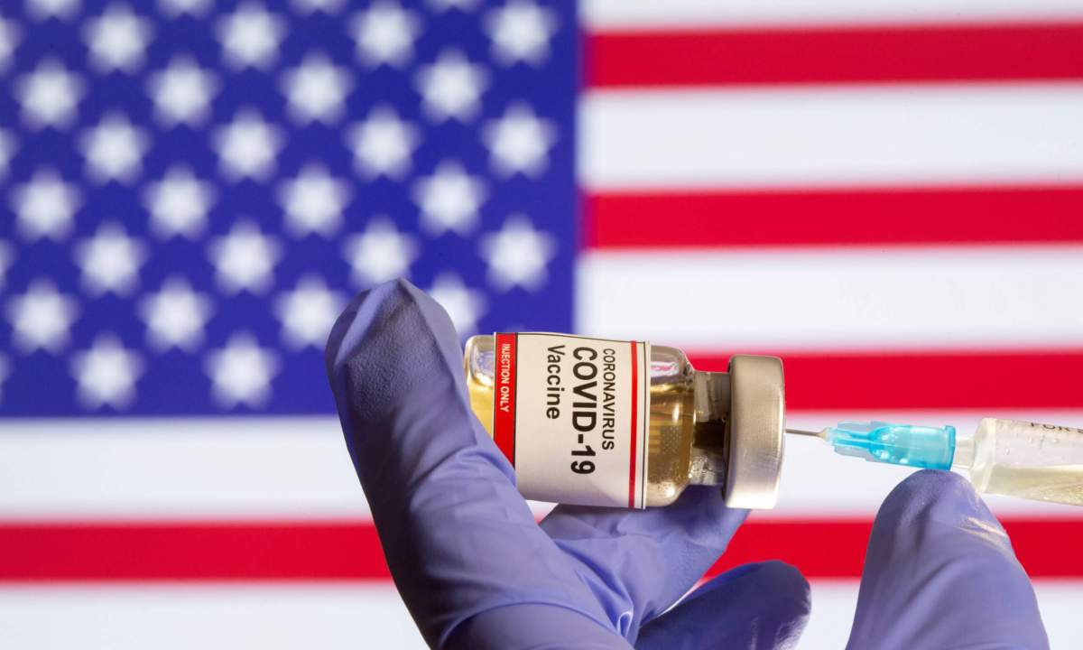 FILE PHOTO: A small bottle labeled with a “Coronavirus COVID-19 Vaccine” sticker and a medical syringe in front of displayed USA flag in this illustration